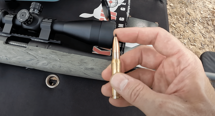 Hand holding a 308 Winchester bullet and a rifle with a scope in the background